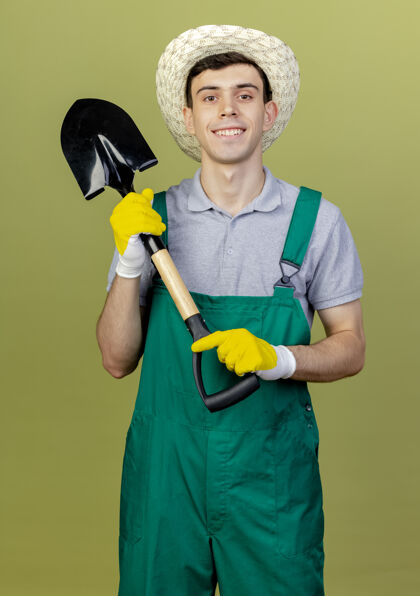 持有微笑着的年轻男园丁戴着园艺帽子和手套拿着铁锹看着相机在橄榄绿的背景上与复制空间隔离园艺复制手套