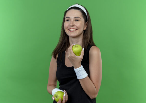 站着戴着头巾的年轻健身女士手持绿色的苹果 站在绿色的背景上 面带幸福的笑容健身头带面子