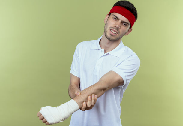 运动戴着头巾的年轻运动型男子站在绿色背景上 摸着缠着绷带的手感到疼痛绷带头带触摸
