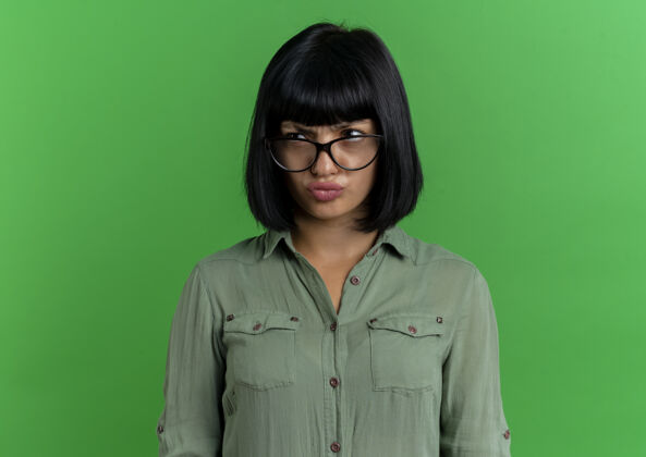 年轻严肃的年轻黑发白种人女孩戴着眼镜看着隔离在绿色背景和复印空间的一面绿色空间黑发