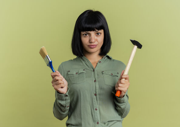 油漆自信的年轻黑发白人女孩拿着画笔和锤子在橄榄绿的背景和复制空间隔离持有年轻锤子