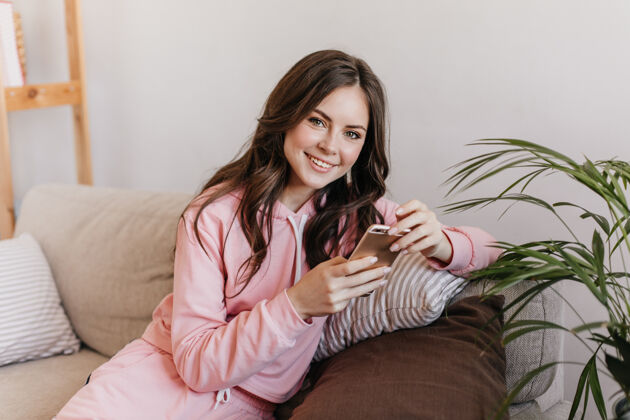 玩家一位身穿粉色连帽衫的黑发女郎正坐在柔软的沙发上 用智能手机观看新照片无聊游戏家庭