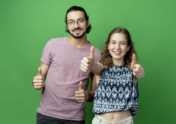微笑一对成功的年轻夫妇看着相机 在绿色的背景下愉快地竖起大拇指成功拇指欢呼