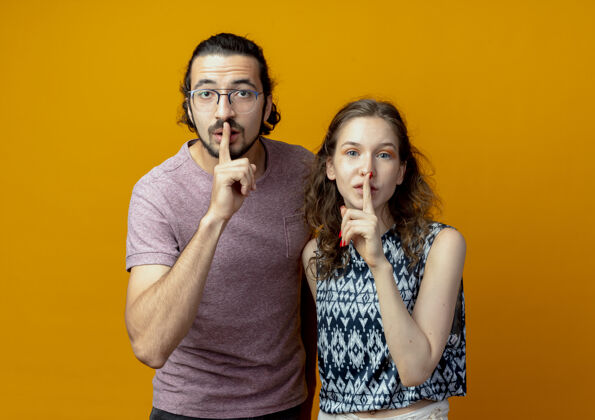 嘴唇一对年轻夫妇看着摄像机 手指放在嘴唇上 站在橙色背景上做着沉默的手势镜头年轻手势