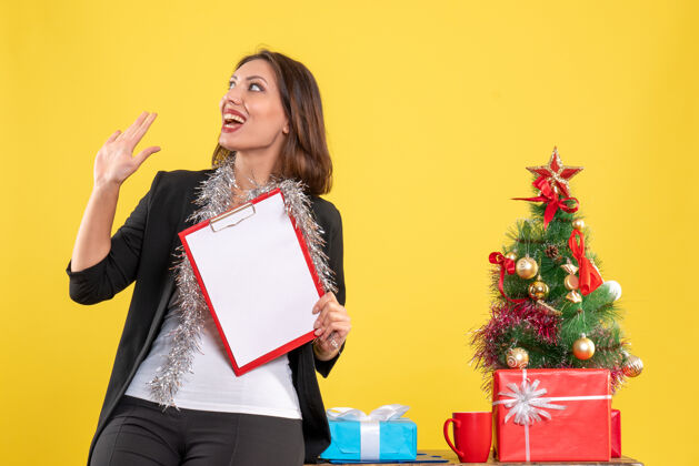 人圣诞气氛与震惊美丽的女士站在办公室里 拿着文件在黄色的办公室举行心情美丽