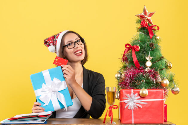秀迷人的女士穿着西装 戴着圣诞老人的帽子和眼镜 在黄色的办公室里展示礼物和银行卡漂亮圣诞老人时尚