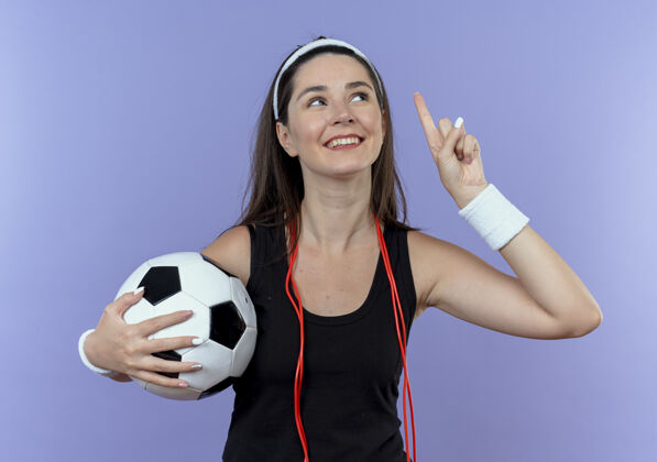 脖子戴着头带 脖子上系着跳绳 手拿足球 手指朝上 面带微笑的年轻健身女士站在蓝色背景上 有了新的想法跳过绳子足球