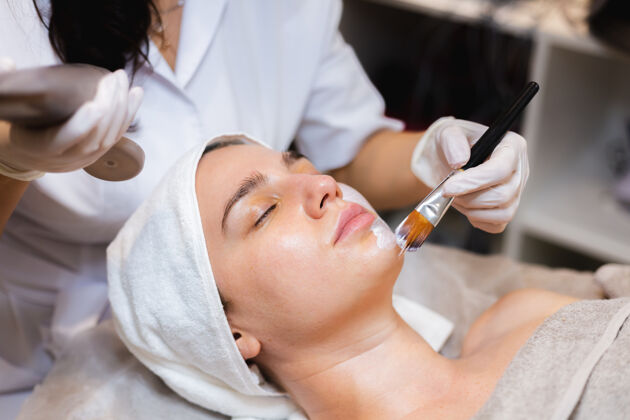 休息美容师用刷子将白色保湿面膜涂在spa美容院一位年轻女客户的脸上放松治疗健康