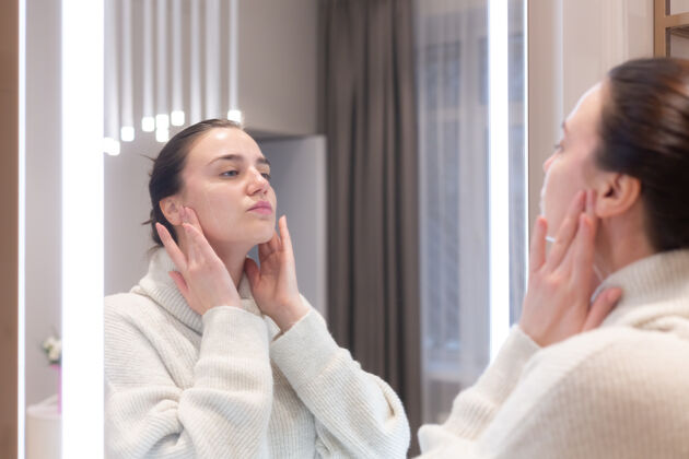 过程在美容院穿毛衣的年轻美女照镜子 抚摸自己的脸 思考即将到来的手术 考虑自己水疗面部健康