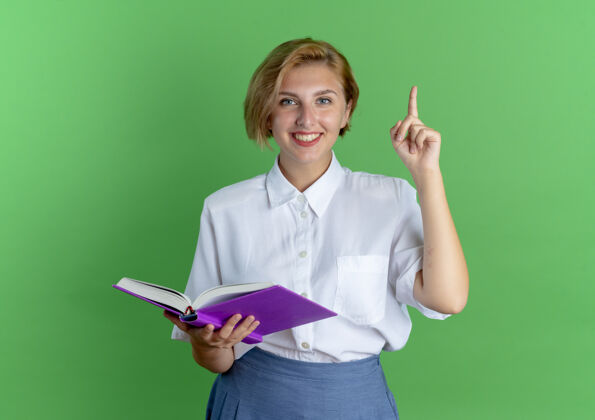 年轻年轻的微笑着的金发俄罗斯女孩拿着一本书 在绿色的背景上孤立地指向上面 还有复印空间微笑指着抱着