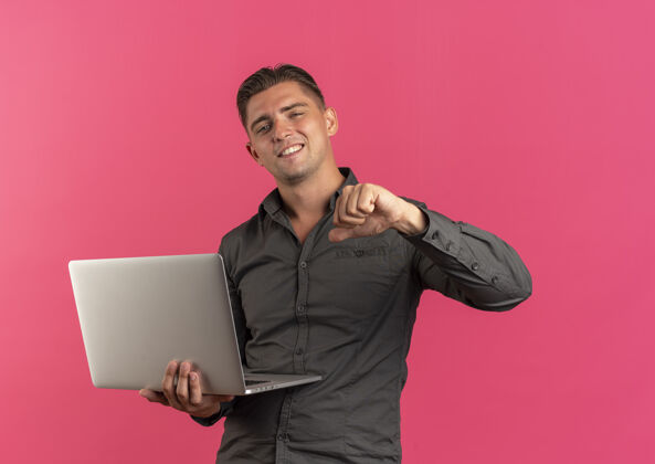 金发年轻自信的金发帅哥拿着笔记本电脑 指着自己被隔离在粉色背景下的复制空间尖自己笔记本电脑