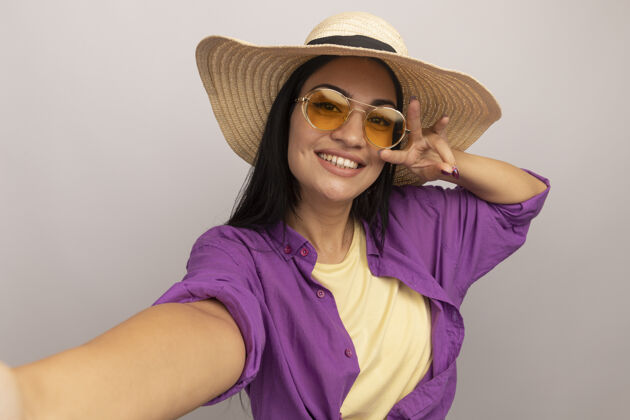 人戴着太阳眼镜 戴着沙滩帽 三个手指的黑发美女微笑着假装站在前面 独自在白墙上自拍衣服帽子感觉