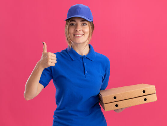 递送身着制服的年轻漂亮的送货员微笑着拿着披萨盒 在粉红色的墙上竖起大拇指站立脸人