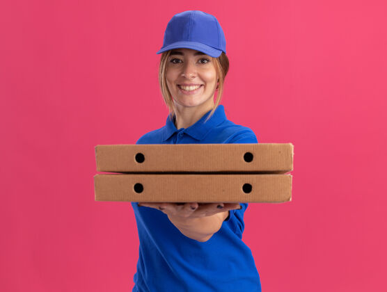 公民身着制服 面带微笑的年轻漂亮女送货员拿着比萨饼盒 看着粉色墙壁上孤立的正面递送人姿势