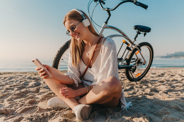 女人时尚年轻迷人的金发微笑的女人坐在沙滩上骑着自行车戴着耳机听音乐海滩热带旅行