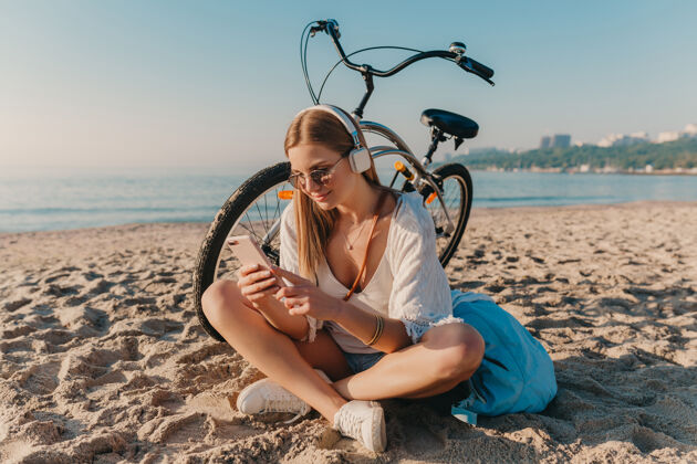 海岸时尚年轻迷人的金发微笑的女人坐在沙滩上骑着自行车戴着耳机听音乐旅程配件耳机