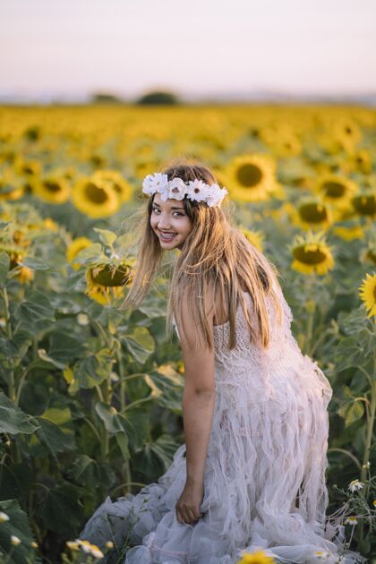 脸美丽的女人穿着白色的连衣裙 微笑着站在向日葵的田野里皮肤头带向日葵