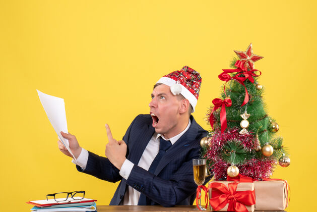 桌子前视图愤怒的男人展示文件坐在圣诞树附近的桌子和黄色背景上的礼物生意人礼物前面