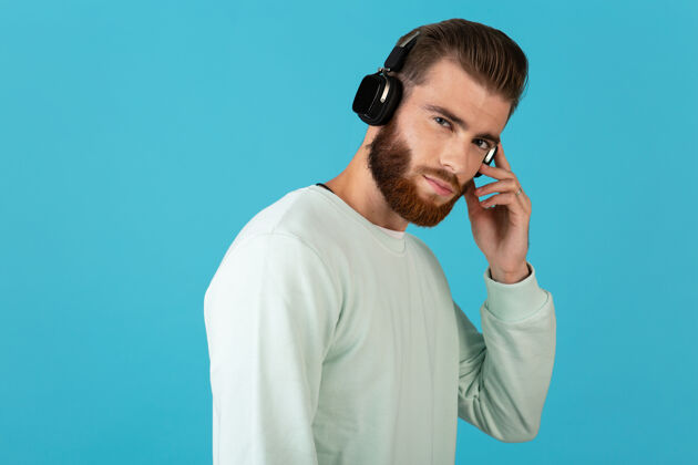 时髦时尚迷人的年轻胡须男子在无线耳机上听音乐现代风格自信的心情隔离在蓝色的墙上时髦立体声声音