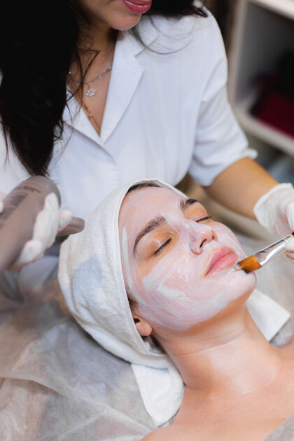 头部美容师用刷子将白色保湿面膜涂在spa美容院一位年轻女客户的脸上放松年轻人