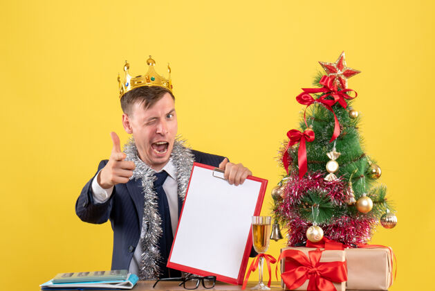 圣诞节前视图：商人眨着眼睛坐在圣诞树旁的桌子旁 呈现黄色背景眼睛礼物前面