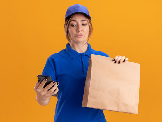 人失望的年轻漂亮的女送货员穿着制服 拿着纸包 看着橙色墙上的电话衣服脸制服