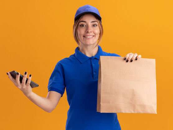 公民身着制服 面带微笑的年轻漂亮女送货员把纸包和电话隔在橙色的墙上包装年轻微笑