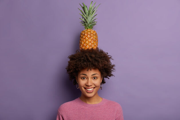 傻瓜一幅迷人的年轻女子的画像 玩弄着成熟的菠萝 看着镜头 笑容灿烂 露出洁白的牙齿 穿着休闲的套头衫牙齿水果有机