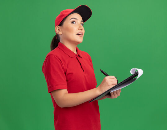 制服身穿红色制服 戴着便笺和铅笔帽子的年轻女送货员站在绿色的墙壁上沉思地抬起头来年轻思考帽子