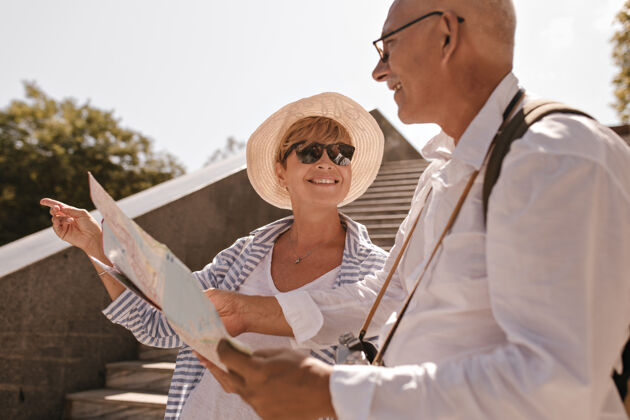 地图带着金色短发 戴着太阳镜 戴着帽子 穿着条纹服装 微笑着的女人指着旁边 看着穿着白衬衫 拿着地图和相机的男人人游客快乐