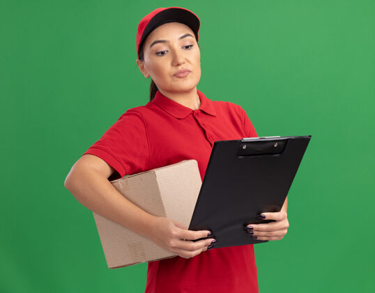 交货年轻的送货员身穿红色制服 戴着帽子 手里拿着纸板箱 手里拿着剪贴板 严肃地站在绿色的墙上看着它纸板立场女人