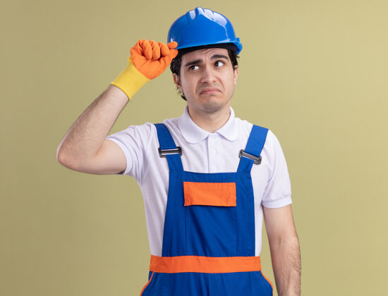 站年轻的建筑工人穿着建筑制服 戴着橡胶手套戴着安全帽 困惑地摸着他站在绿墙上的头盔制服安全建筑工人