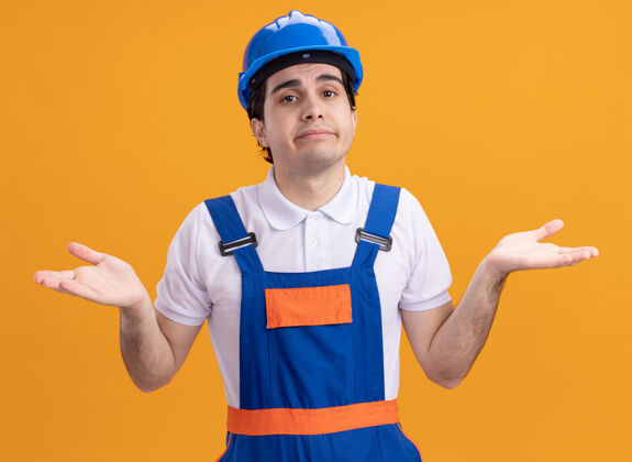 站立年轻的建筑工人穿着建筑制服 戴着安全帽 困惑地看着前面 双臂伸向两边 站在橙色的墙上 没有回答头盔建设者伸展