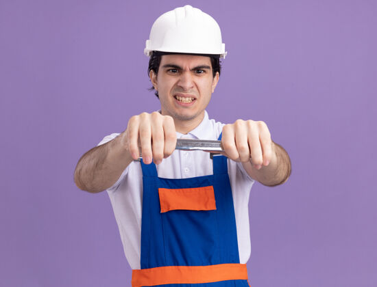 头盔身穿施工制服 头戴安全帽 手持扳手的年轻建筑工人站在紫色的墙上怒气冲冲地看着它男人站着愤怒