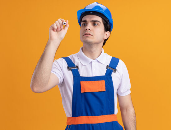 写字身穿施工制服 头戴安全帽的年轻建筑工人站在橙色的墙上 脸上带着严肃的表情 在前面用笔写字制服前面安全