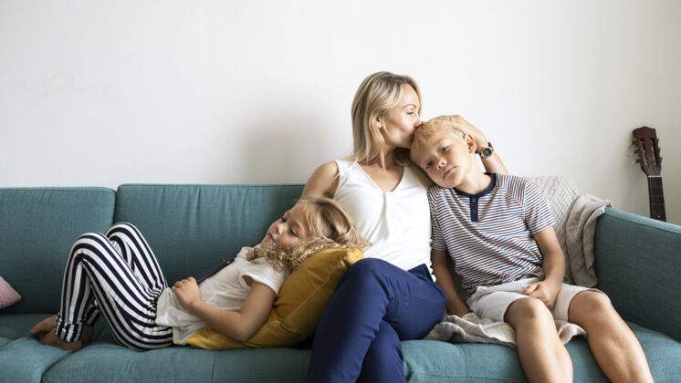 母亲金发妈妈亲吻儿子的头 和女儿在沙发上放松年轻男孩儿子