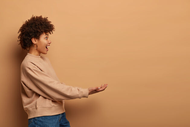 高兴一个快乐的卷曲的非裔美国女人的侧面照片假装拿着什么东西 伸出双手 穿着宽松的棕色套头衫和牛仔裤 站在米色的墙上喜悦携带无形