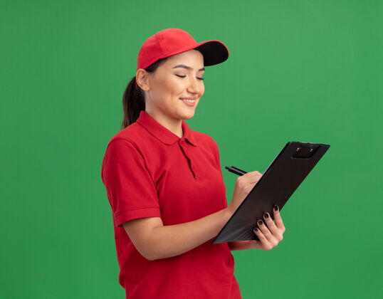 年轻身穿红色制服 头戴便帽 拿着写字板和铅笔的年轻女送货员站在绿色的墙边愉快地微笑着微笑剪贴板站立