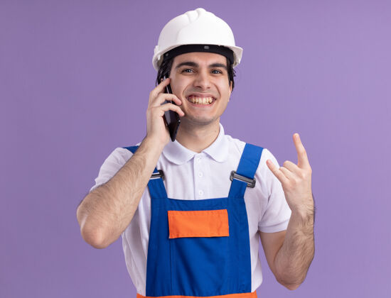 头盔身穿施工制服 头戴安全帽的年轻建筑工人站在紫色的墙上 兴高采烈地微笑着用手机制作岩石符号手机岩石站立