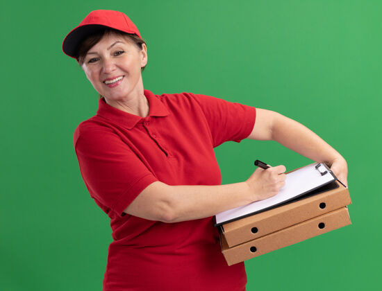 空白身穿红色制服 头戴鸭舌帽的中年女送货员 手里拿着披萨盒和空白页的剪贴板 用笔写字 面带微笑地站在绿色的墙上 看着前面页面帽子剪贴板