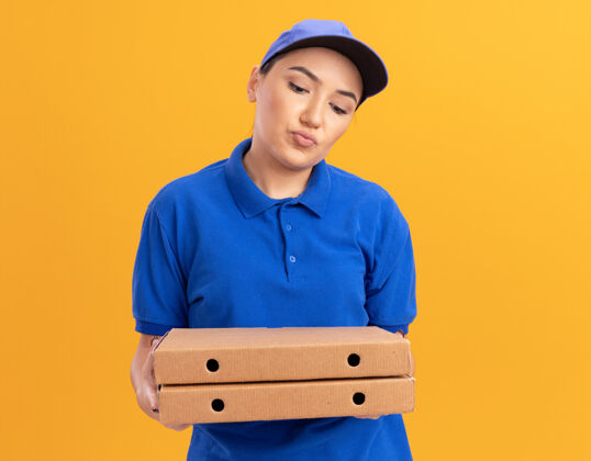 焦虑身穿蓝色制服 头戴鸭舌帽 拿着比萨饼盒的年轻女送货员站在橘色的墙上 看着这些比萨饼盒感到困惑和焦虑站着披萨年轻