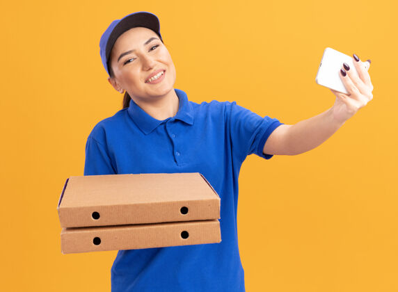 自拍身穿蓝色制服 戴着帽子 拿着披萨盒 用智能手机自拍的年轻女送货员站在橙色的墙上开心地微笑着披萨制服送货