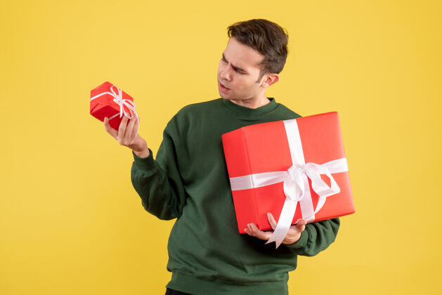 人物前视图年轻人与圣诞礼物检查他的礼物站在黄色背景上手持专业正面