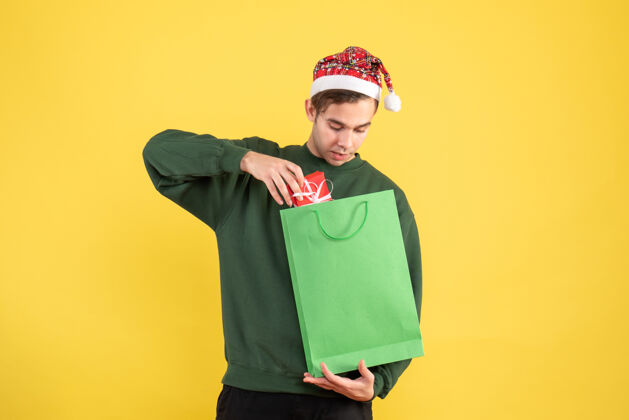男人正面图戴着圣诞帽的年轻人拿着绿色的购物袋和礼物站在黄色的背景复制空间绿色复制年轻人