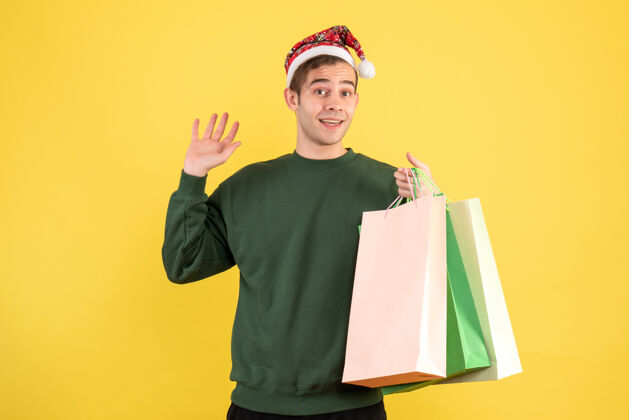 成人正面图：戴着圣诞帽的年轻人拿着购物袋站在黄色背景上帽子前面工作