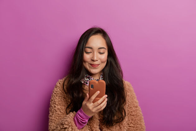 女性快乐的亚洲年轻女子 黑发 看有趣的新闻 有小玩意瘾 很高兴收到信息 穿着棕色外套外套手机发送