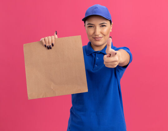 递送快乐的年轻送货员身穿蓝色制服 戴着帽子 拿着纸包 食指指着前面 微笑着自信地站在粉红色的墙上女人纸制服