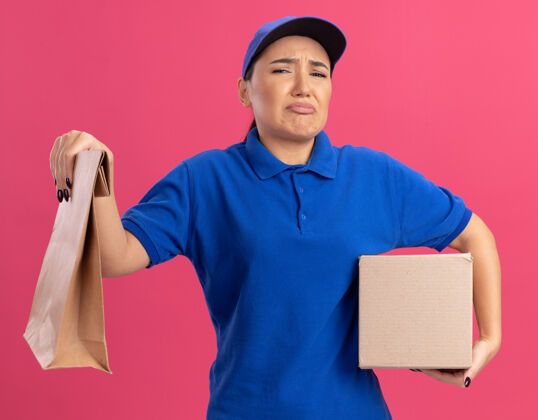 包装一个穿着蓝色制服 戴着帽子 拿着纸包和纸箱的年轻送货员站在粉红色的墙上 愁眉苦脸地看着前面盒子站着送货