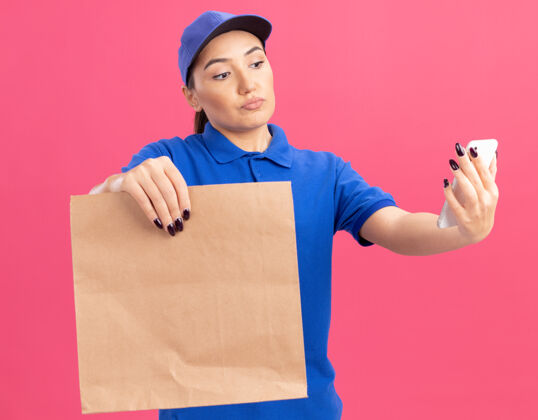 帽子身穿蓝色制服 戴着帽子的年轻送货员拿着纸包 站在粉红色的墙上 严肃地看着自己的智能手机递送脸严肃