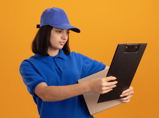 脸身穿蓝色制服 头戴鸭舌帽的年轻送货女孩拿着纸板箱和剪贴板 严肃地站在橙色的墙上看着它立场剪贴板交货